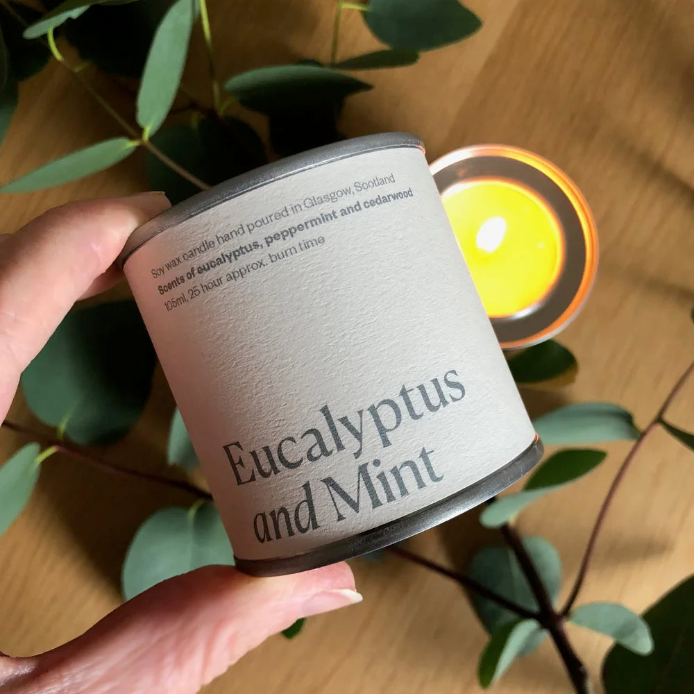 Candle labeled 'Eucalyptus and Mint' held by a hand with a blurred candle in the background.