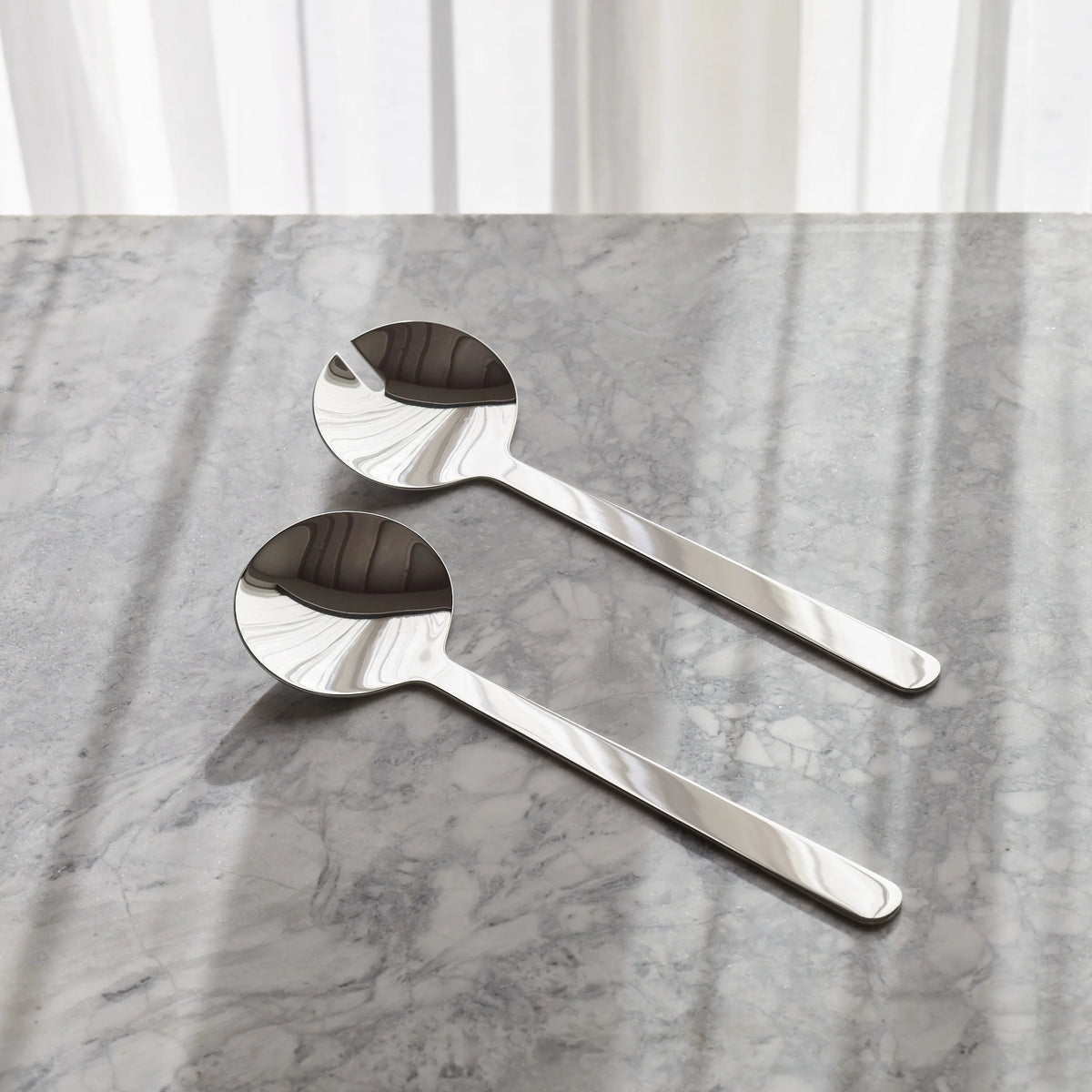 Two polished stainless steel spoons with a flared, textured bowl, resting on a gray marble surface with soft window light shadows.