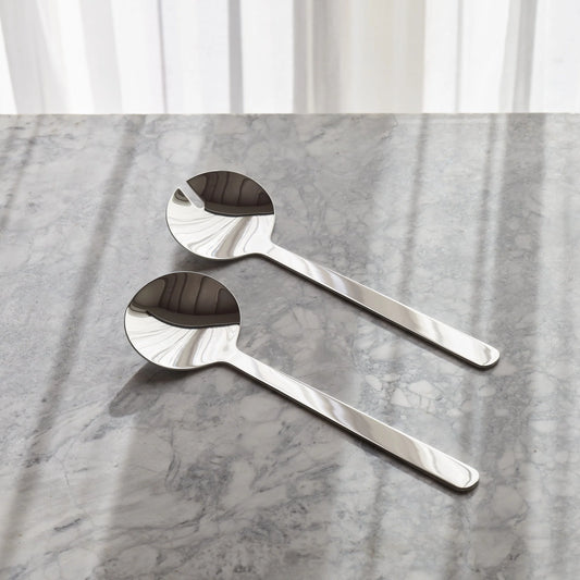 Two polished stainless steel spoons with a flared, textured bowl, resting on a gray marble surface with soft window light shadows.