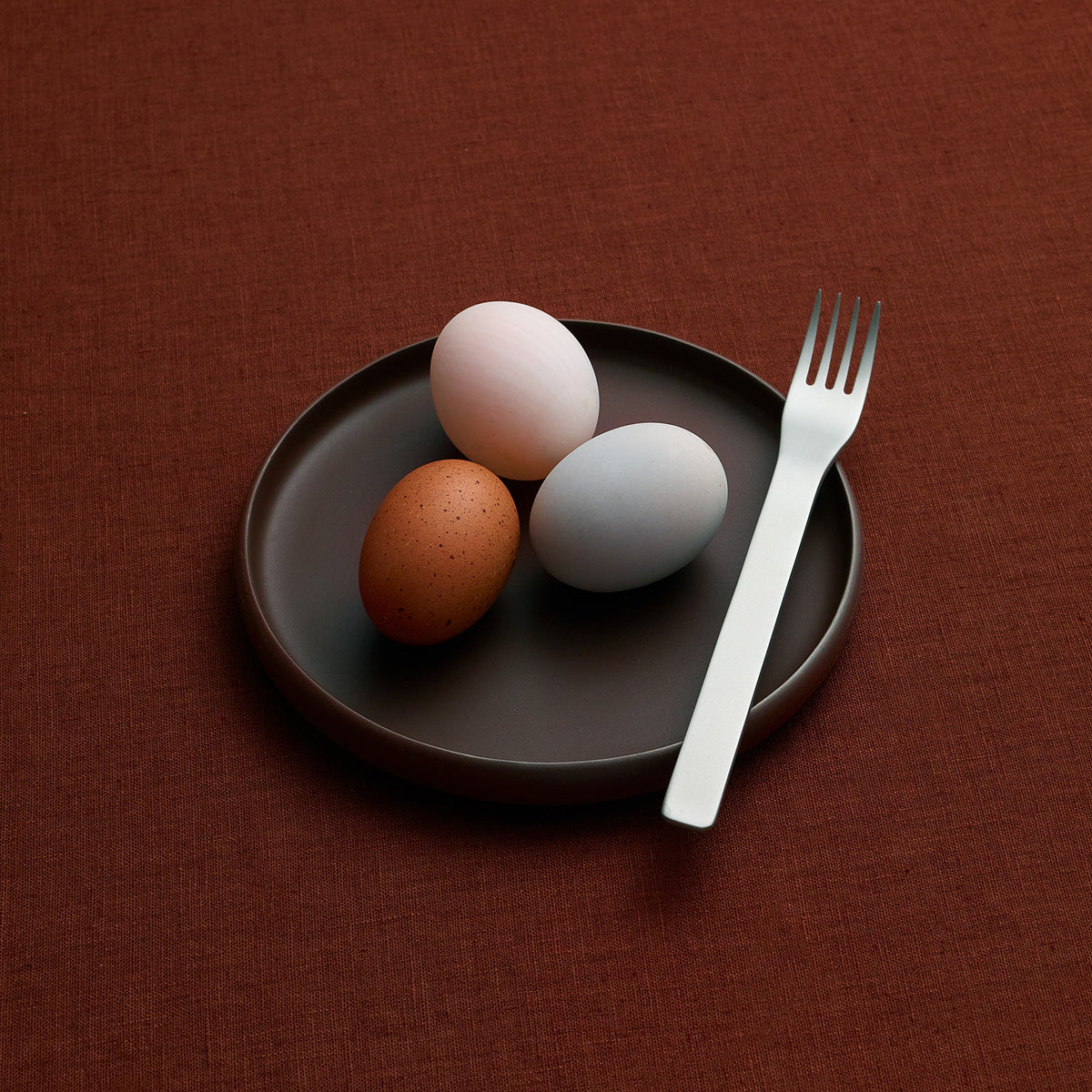 Three eggs in different colors resting on a dark round plate next to a white plastic fork, all set on a textured dark brown surface.