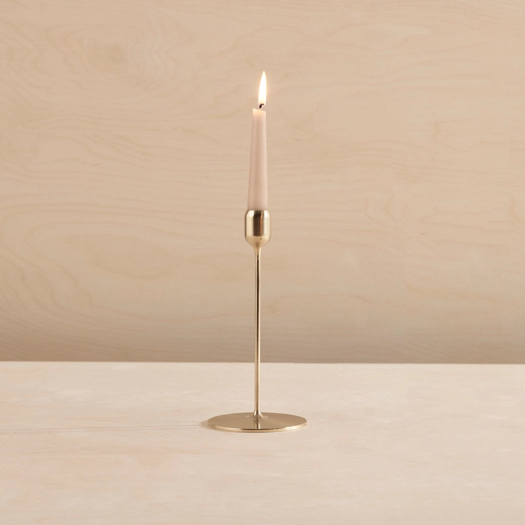 A single lit taper candle in a slim brass holder with a round base, standing on a light wood surface against a pale wood background.