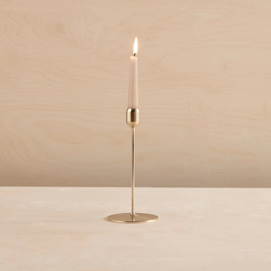 A single lit taper candle in a slim brass holder with a round base, standing on a light wood surface against a pale wood background.