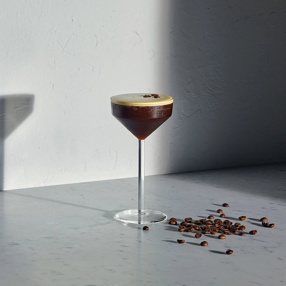 A martini-style cocktail filled with dark coffee and topped with a light foam, served in a stemmed coupe glass on a marble surface with scattered coffee beans against a textured white wall.