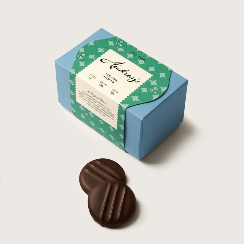 A blue and green patterned box of Achingly chocolate truffles with two dark chocolate mints placed in front, on a light beige background.