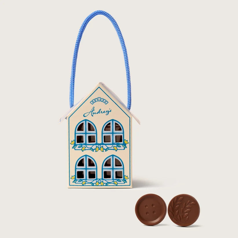 A small house-shaped chocolate gift box with a blue rope handle, decorated with blue window frames and floral details, alongside two round milk chocolate buttons on a light beige background.
