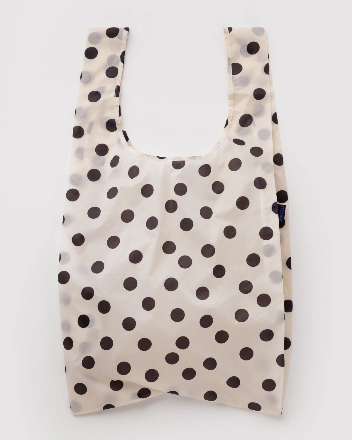 A reusable plastic shopping bag with a black polka dot pattern on a warm off-white background, shown against a plain white studio background.