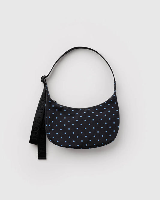 A crescent-shaped shoulder bag in black with a light blue polka dot pattern, featuring a black adjustable strap with white lettering, photographed on a light gray background.