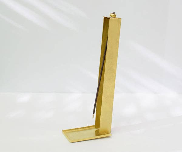 A brass incense holder with a vertical rod holding a dark incense stick, set against a white background with soft leaf shadows.
