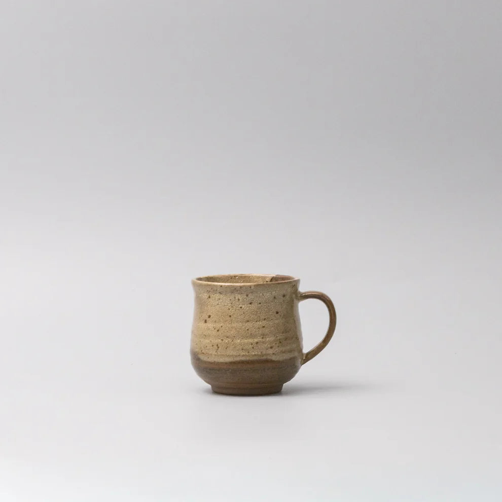 A small handmade ceramic mug with a textured, speckled glaze in earthy beige and brown tones, featuring a simple C-shaped handle, set against a plain light gray background.