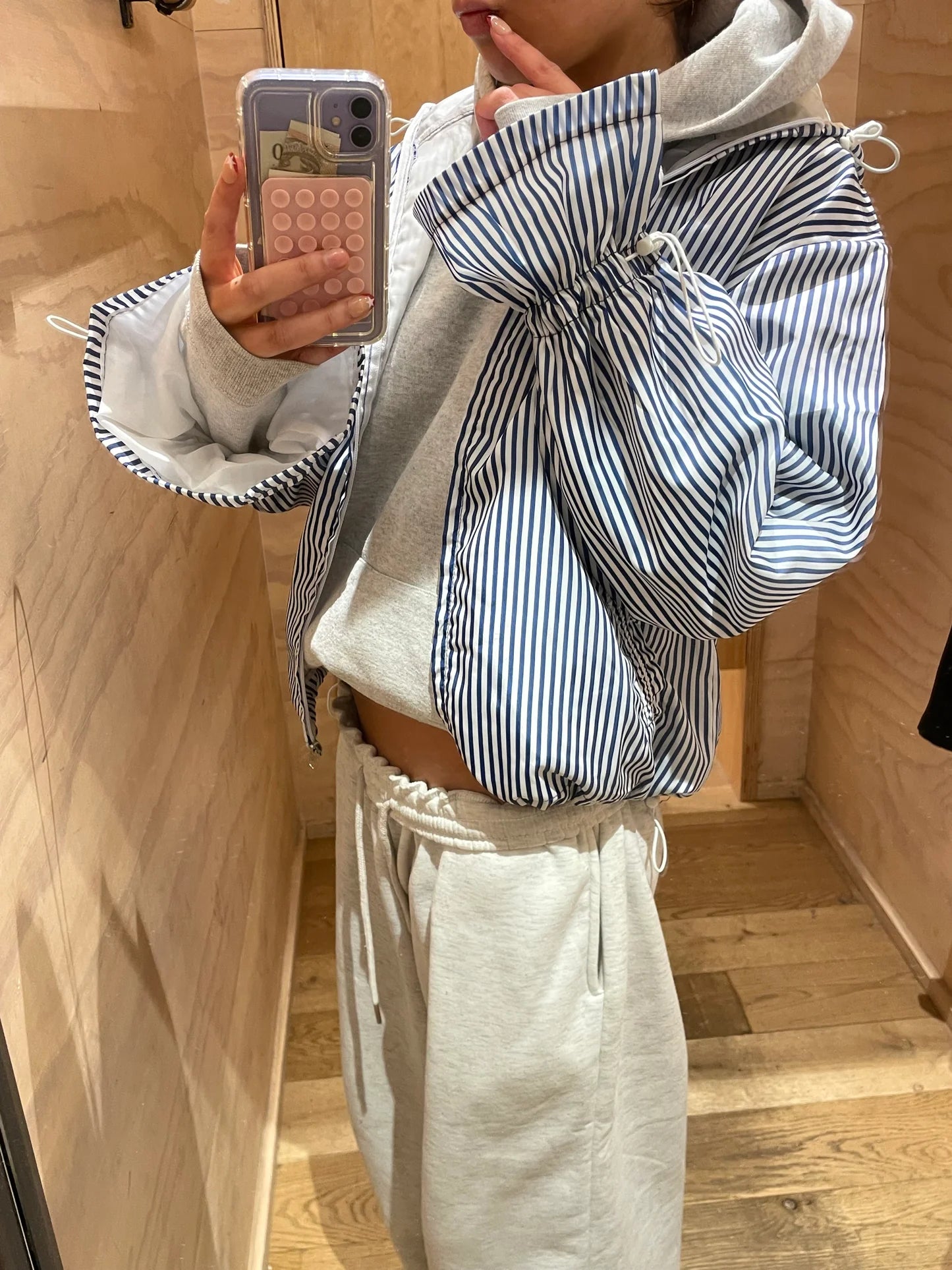Person wearing a gray hoodie with striped sleeves jacket and white pants in a wooden interior setting.