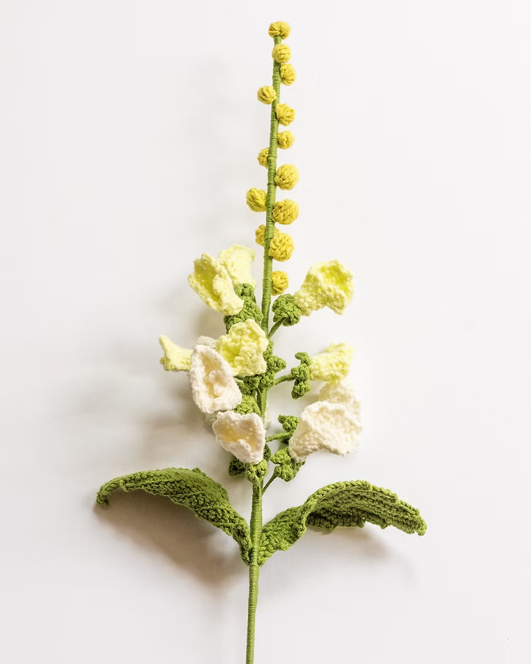 A crocheted artificial flower stem featuring pale yellow and off-white blossoms with textured green leaves, displayed against a clean white background.