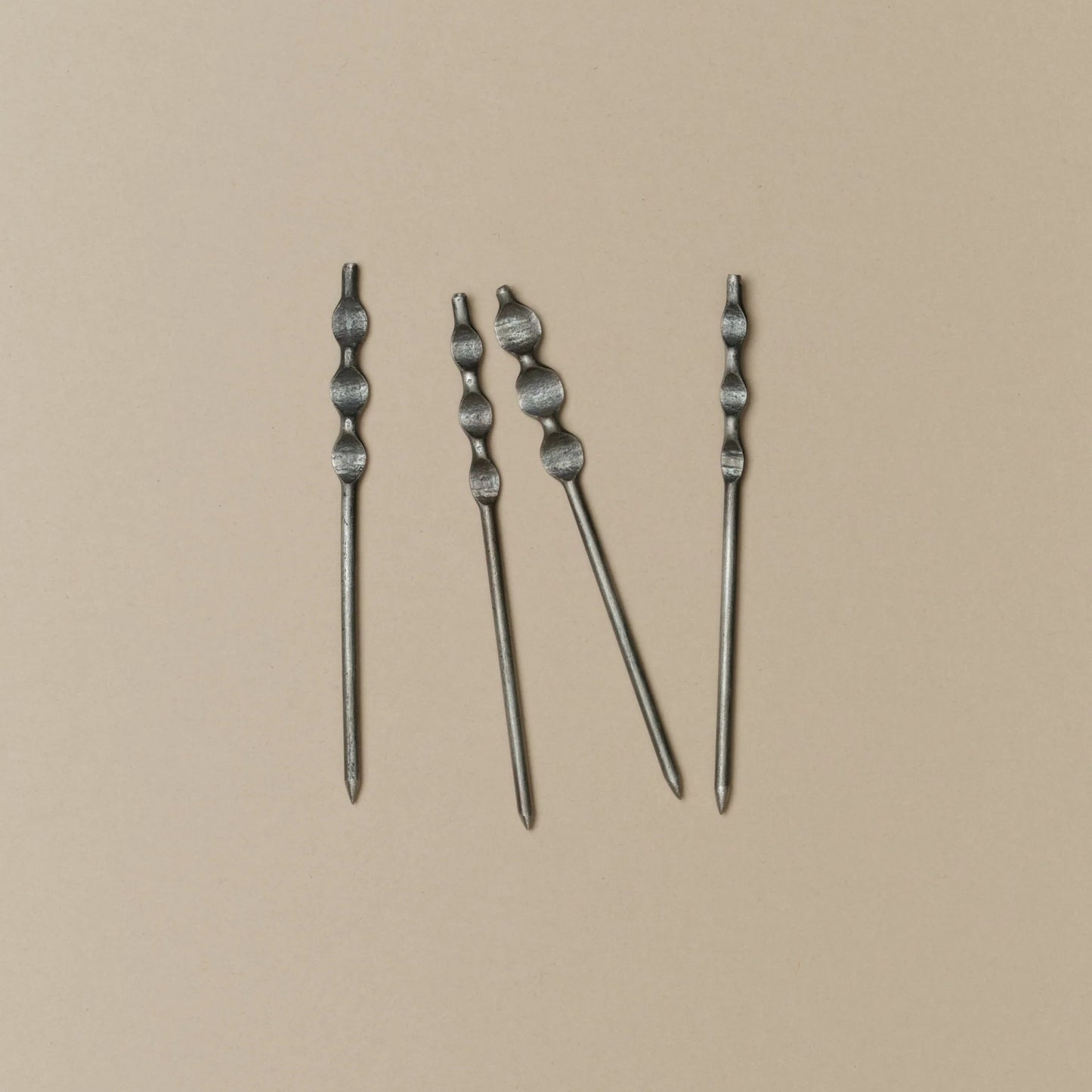 Four silver-toned metal cockail skewers with textured, knotted tops arranged vertically on a solid beige background.