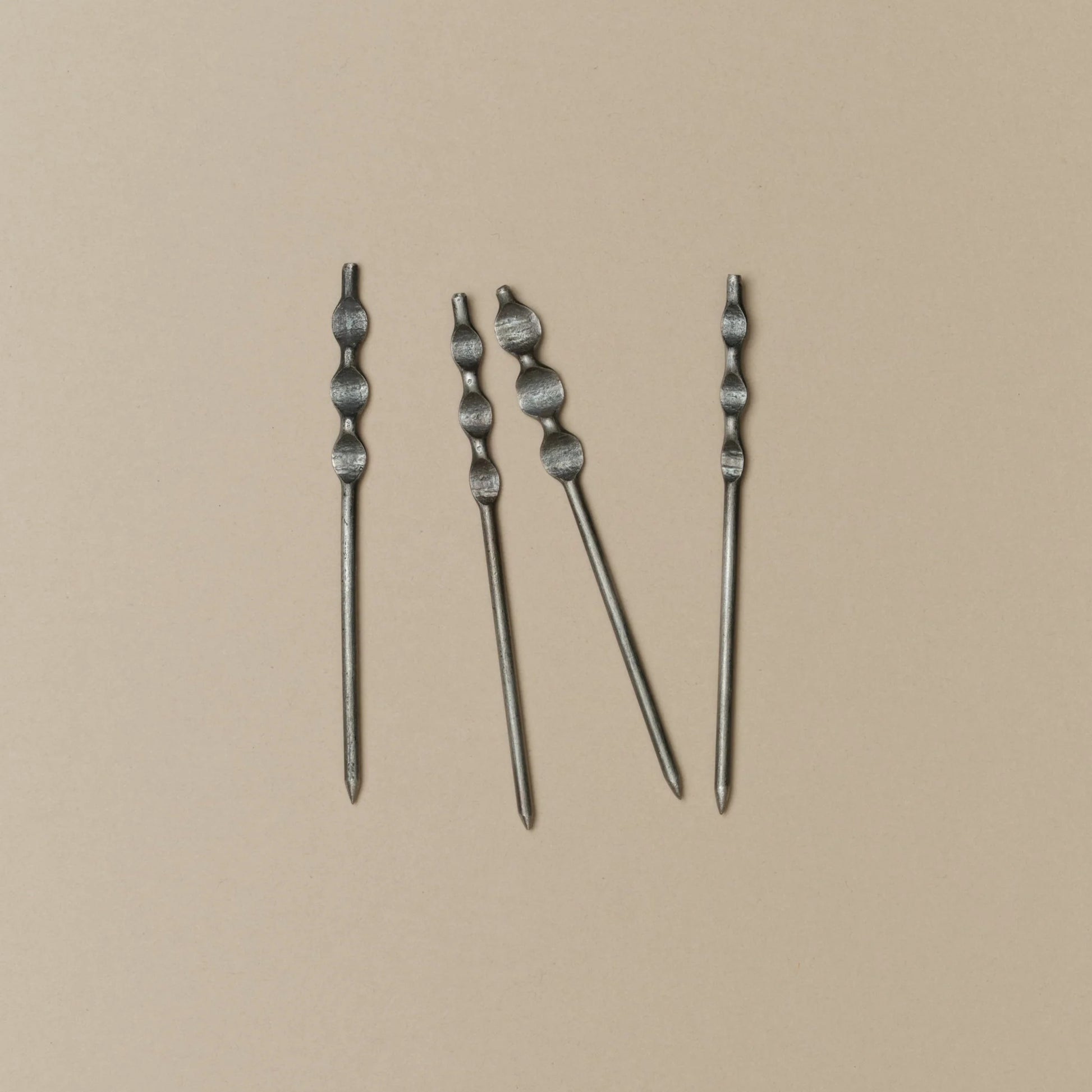 Four silver-toned metal cockail skewers with textured, knotted tops arranged vertically on a solid beige background.