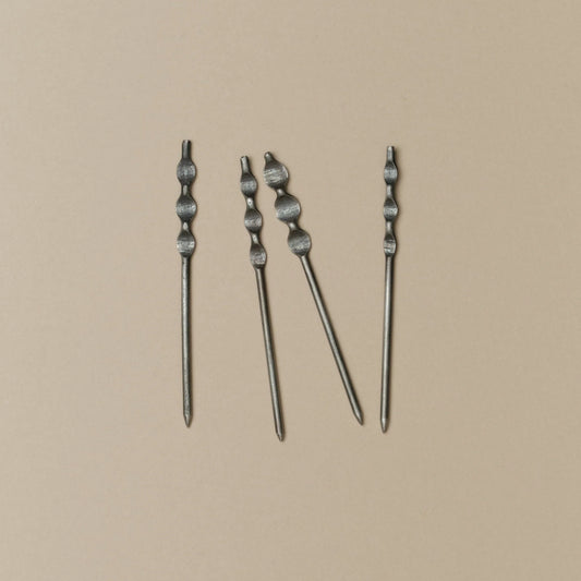 Four silver-toned metal cockail skewers with textured, knotted tops arranged vertically on a solid beige background.