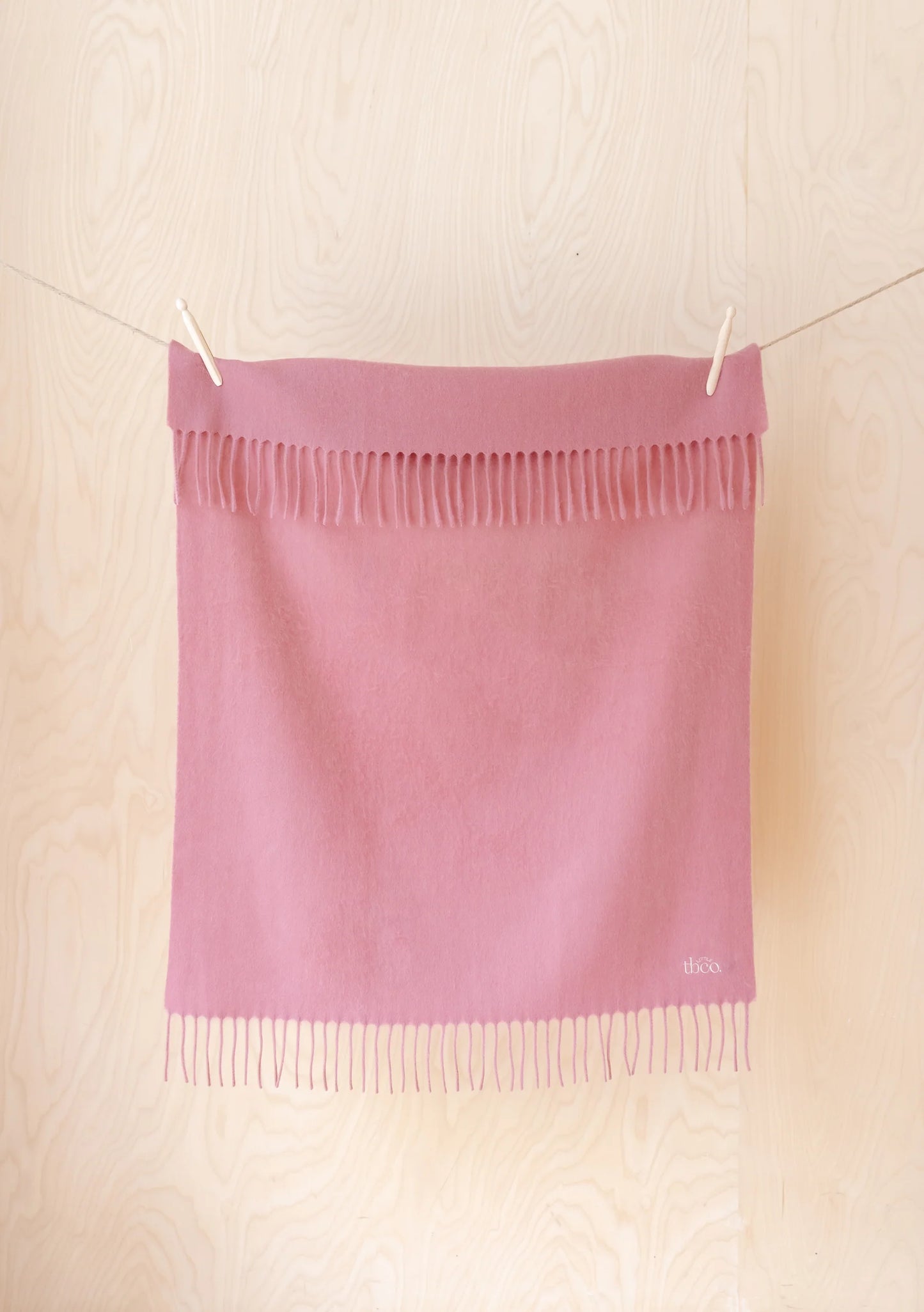 A dusty pink woven scarf with fringe trim, hanging from a string with wooden clothespins against a light wood-paneled wall. 