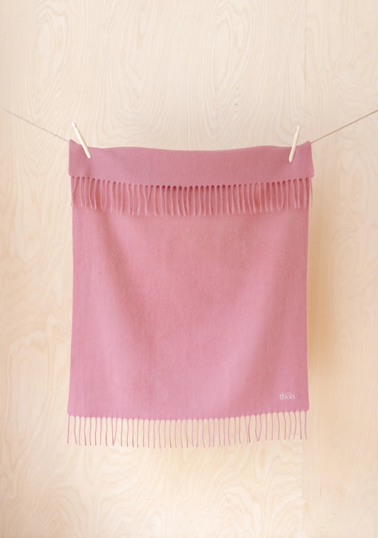 A dusty pink woven scarf with fringe trim, hanging from a string with wooden clothespins against a light wood-paneled wall. 