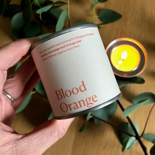 Hand holding a 'Blood Orange' candle with a lit candle in the background