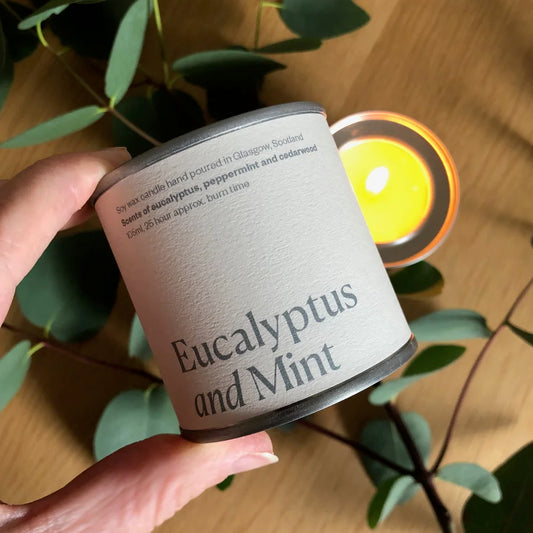 Candle labeled 'Eucalyptus and Mint' held by a hand with a blurred candle in the background.