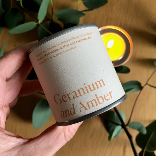 Candle labeled 'Geranium and Amber' held by a hand with a blurred candle in the background.