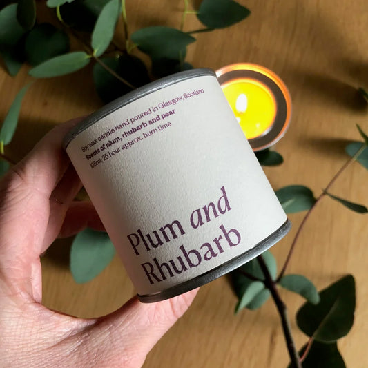 Hand holding a candle labeled 'Plum and Rhubarb' with a blurred candle in the background.