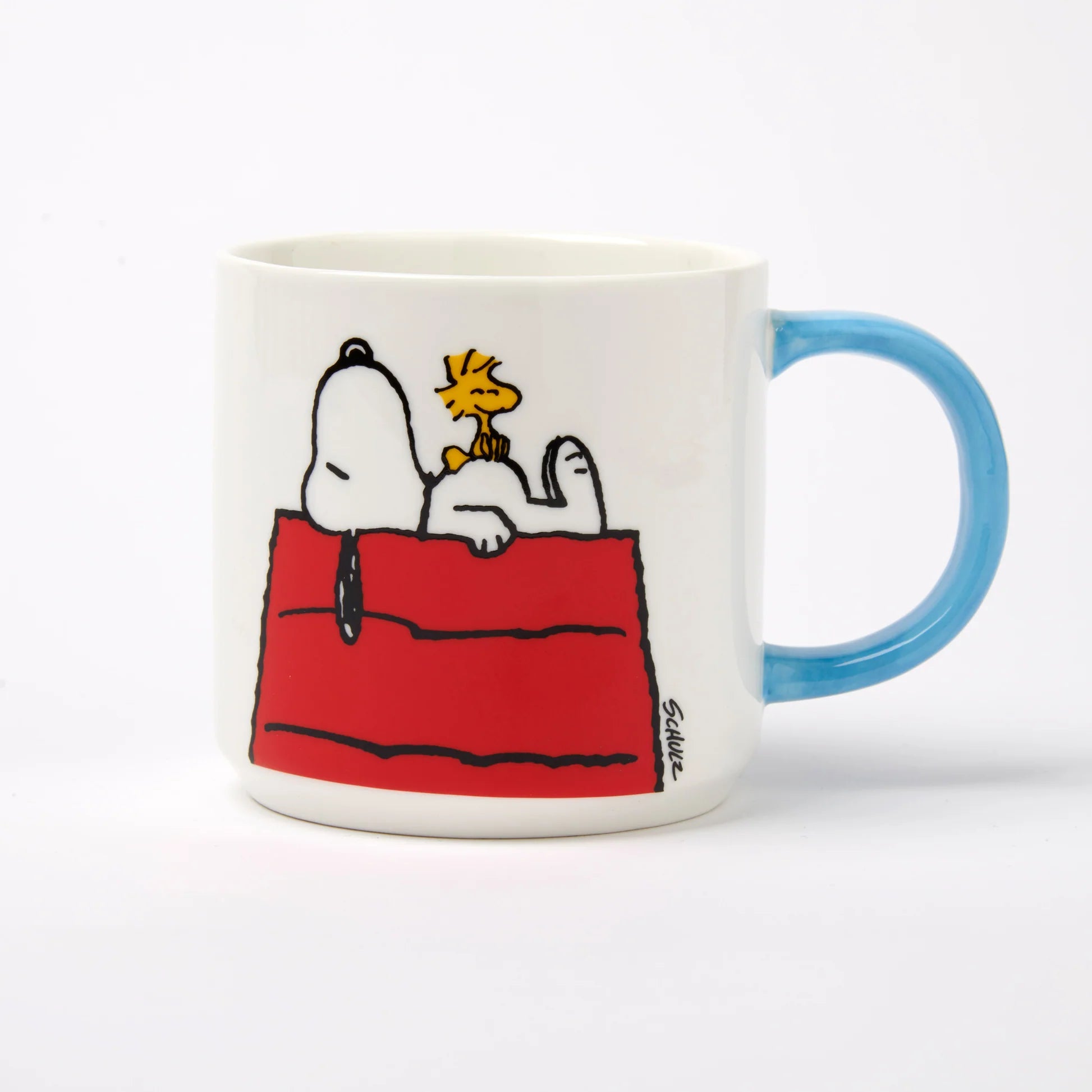 A white ceramic coffee mug featuring a cartoon illustration of Snoopy and Woodstock on a red dog bed, with a light blue handle, photographed on a clean white background.