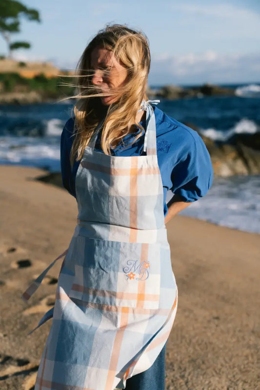 A woman standing on a sandy beach wearing a light blue and white plaid apron with a monogrammed pocket over a blue shirt, with the ocean and rocks in the background.