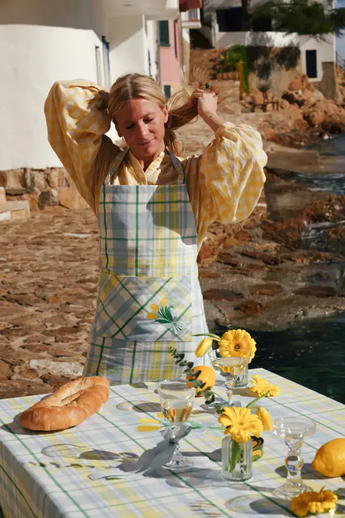Woman in a yellow dress with a floral apron standing by a table outdoors.