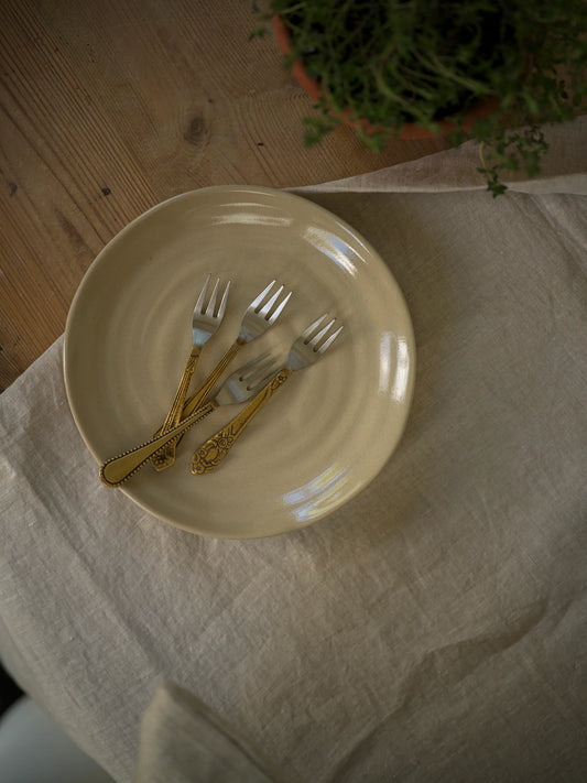 Set of gold forks on a beige plate with a white cloth and plant in the background