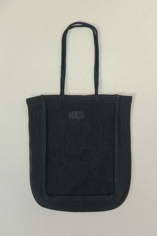 A black woven tote bag with long shoulder straps, featuring a small rectangular metal brand plaque on the front, set against a plain light gray background.