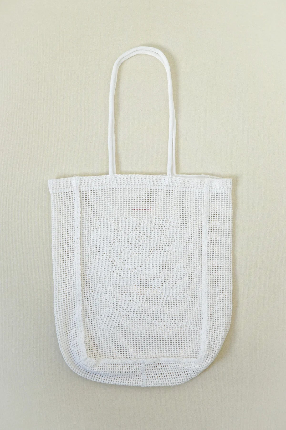 A white open crochet tote bag with long handles, shown flat against a light beige background.