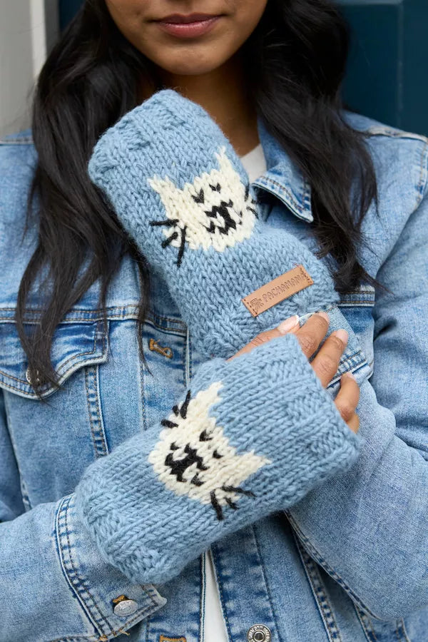 A close-up of a person wearing light blue knit fingerless gloves with white cat faces and black whisker details, styled over a light blue denim jacket.