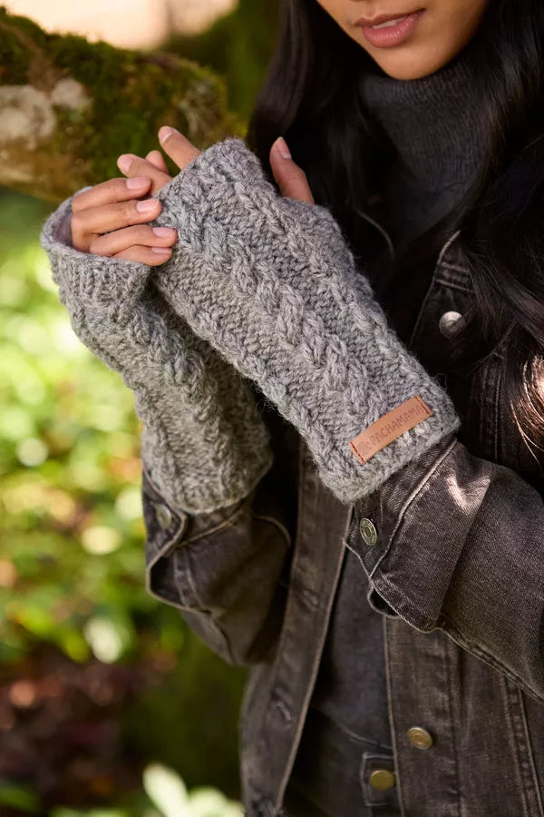 A close-up of a person wearing chunky gray cable-knit fingerless gloves with a small tan leather brand tab, styled with a dark gray turtleneck and distressed denim jacket against a blurred outdoor background.