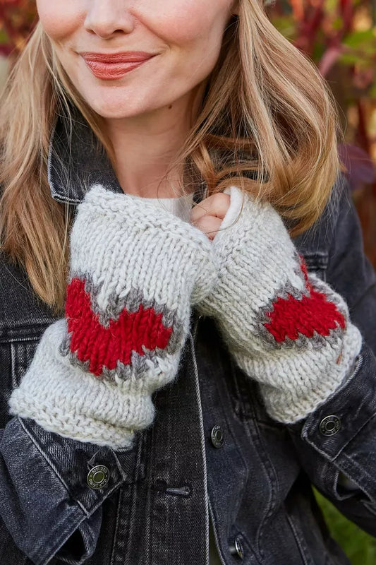Close-up of a woman wearing chunky knit fingerless gloves in off-white with red and gray heart patterns, paired with a dark denim jacket.