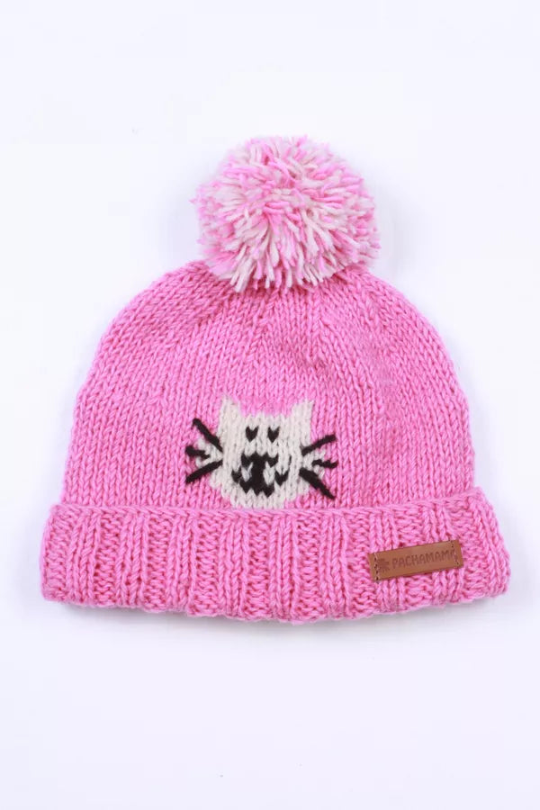 A pink knit beanie hat with a fluffy pom-pom on top, featuring a white and black cat face motif on the front and a small brown leather brand patch on the cuff.