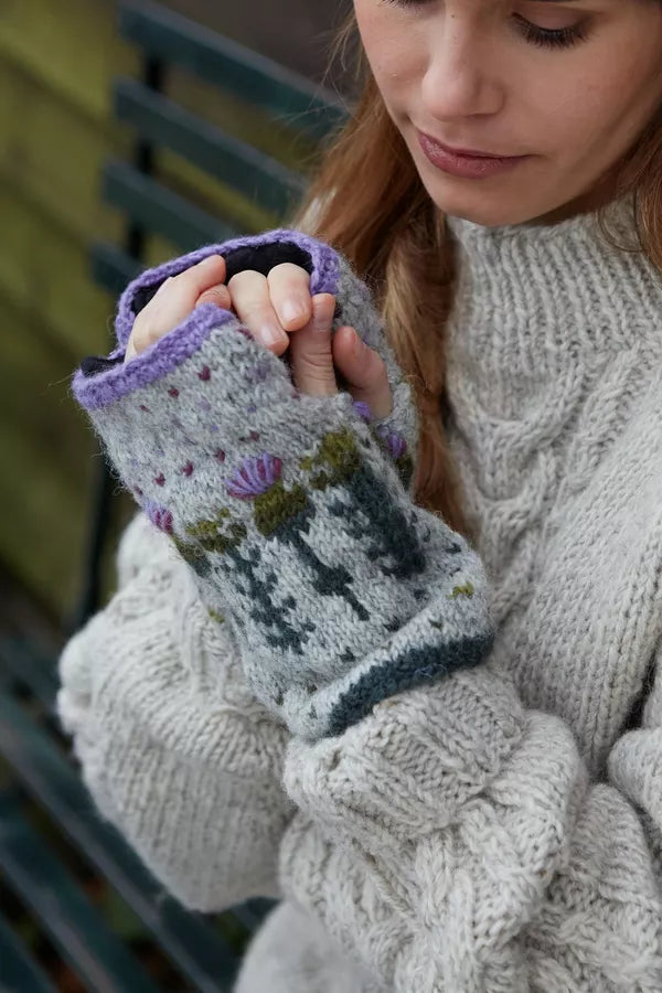 A close-up of a woman wearing a chunky cream cable-knit sweater and purple fingerless knit gloves.