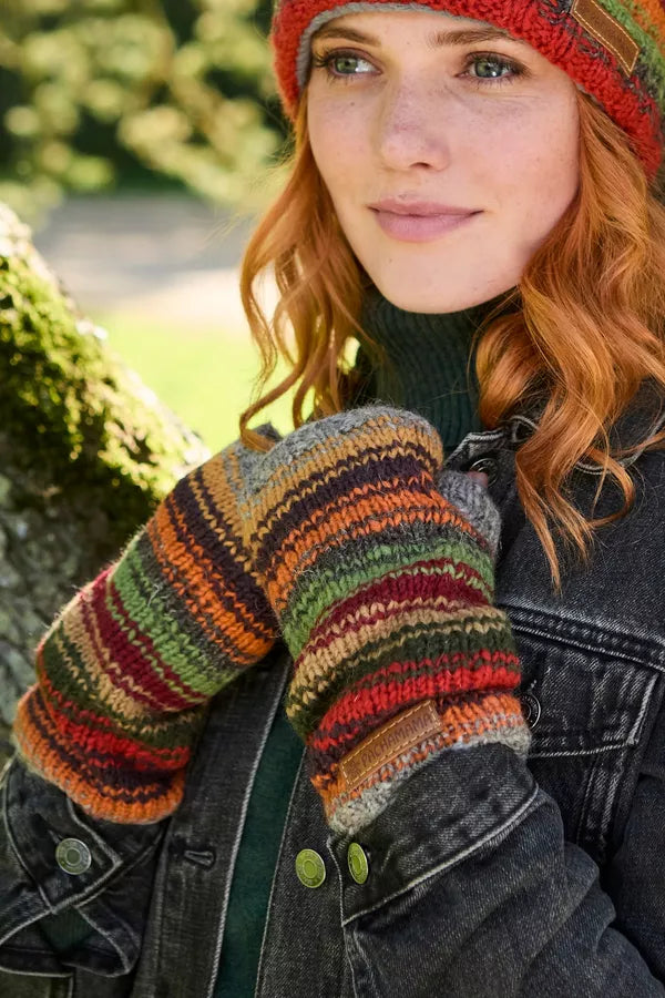A close-up lifestyle shot of a woman outdoors wearing a colorful striped knit beanie, matching knit mittens, and a dark denim jacket.