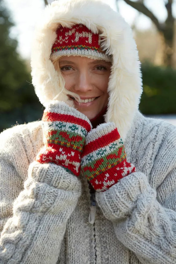 A smiling woman outdoors wearing a white faux-fur-lined knit hooded jacket, cream cable-knit sweater, red knit hat, and red knit mittens with green and yellow pattern details.
