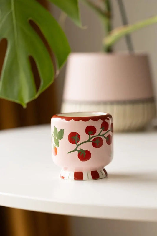 Decorative ceramic cup with tomato design on a white surface, with a blurred plant and lamp in the background.