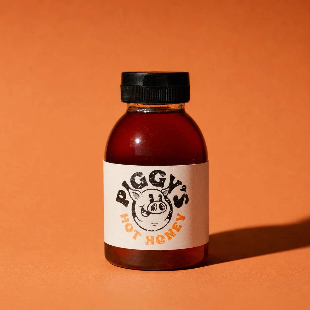 A small plastic bottle of Piggy's Hot Honey with a black cap and a cream label featuring a pig illustration, set against a solid orange background.