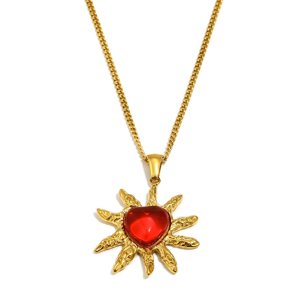 A gold-toned necklace featuring a pendant shaped like a sunburst with a faceted red heart gemstone in the center, shown against a plain white background.