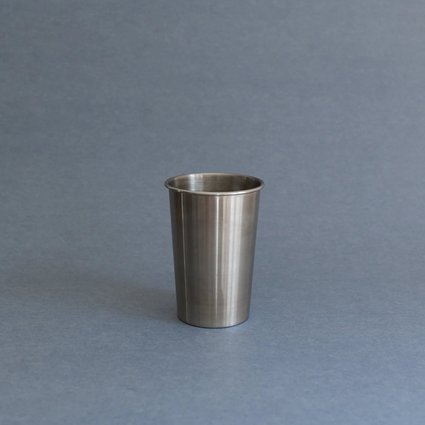 A brushed stainless steel cup or tumbler with a flared rim, standing upright on a smooth gray surface against a matching gray background.
