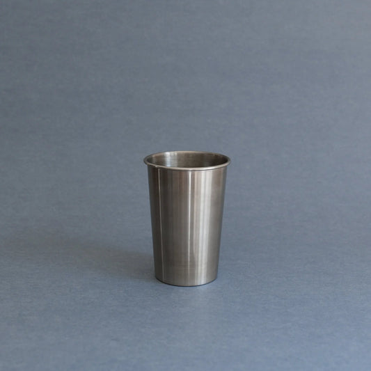 A brushed stainless steel cup or tumbler with a flared rim, standing upright on a smooth gray surface against a matching gray background.
