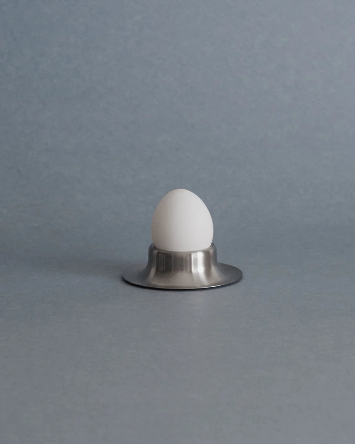 A white ceramic egg-shaped object displayed upright in a small brushed silver metal holder against a smooth gray background.