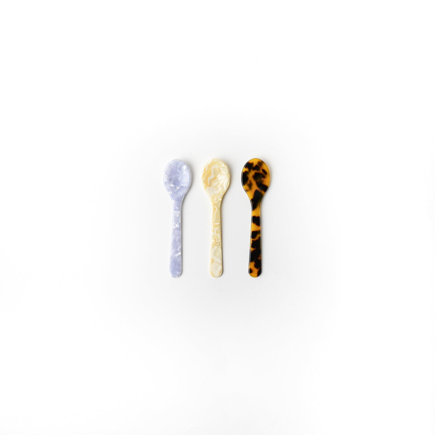 Three small decorative spoons arranged vertically on a plain white background. The left spoon is light lavender with a marbled pattern, the middle spoon is cream with a marbled pattern, and the right spoon is tortoiseshell in brown and black.