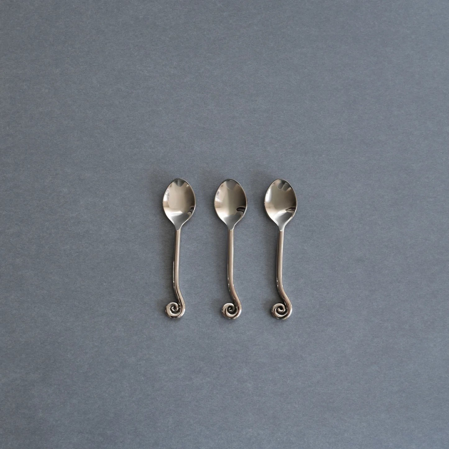 Three small silver spoons with decorative curled handles, arranged side by side on a textured gray background.