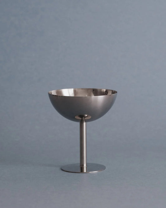 A polished silver metal stemware piece with a shallow bowl and a single cylindrical stem, standing on a round base against a solid blue-gray background.