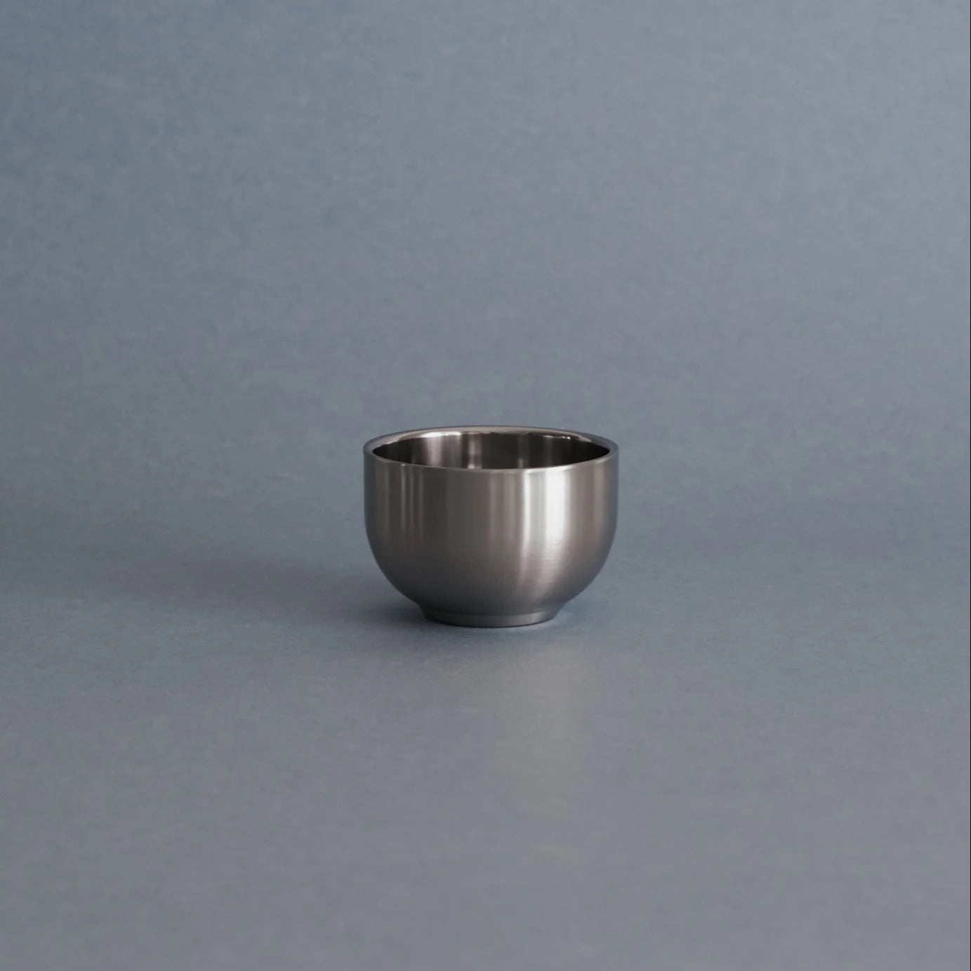 A small, round stainless steel cup or bowl with a brushed metallic finish, centered on a smooth, solid gray surface with a matching gray background.