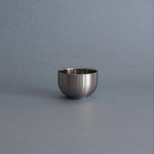 A small, round stainless steel cup or bowl with a brushed metallic finish, centered on a smooth, solid gray surface with a matching gray background.