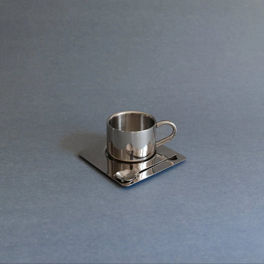 A polished silver metal cup with a handle, resting on a matching square saucer with a small spoon, set against a solid gray background.
