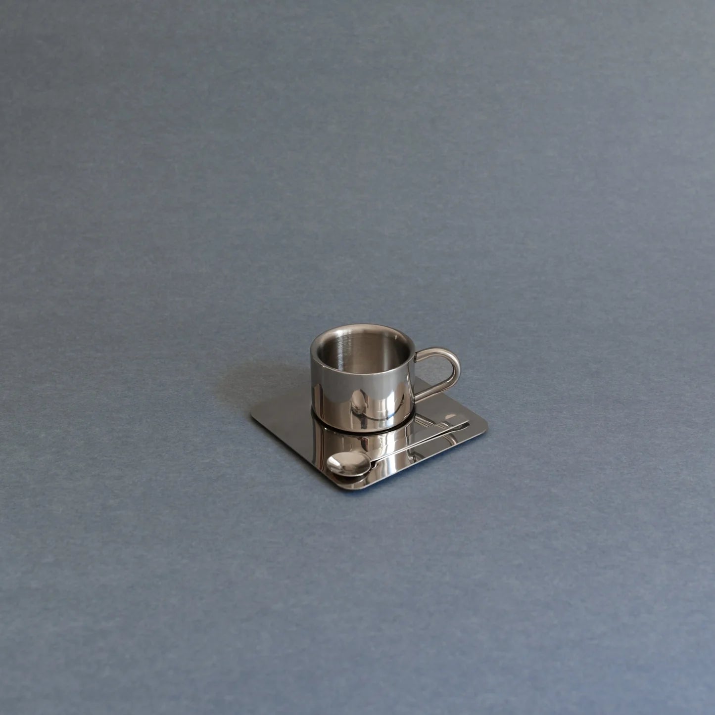 A small metallic silver coffee cup-shaped object resting on a square matching base with a spoon detail, photographed on a plain gray background.
